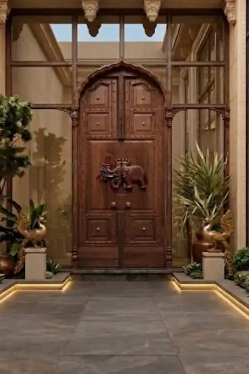 Boutique Couture Grand entryway with a tall, ornate wooden door featuring an elephant relief, flanked by glass panels and potted plants in a luxurious foyer.