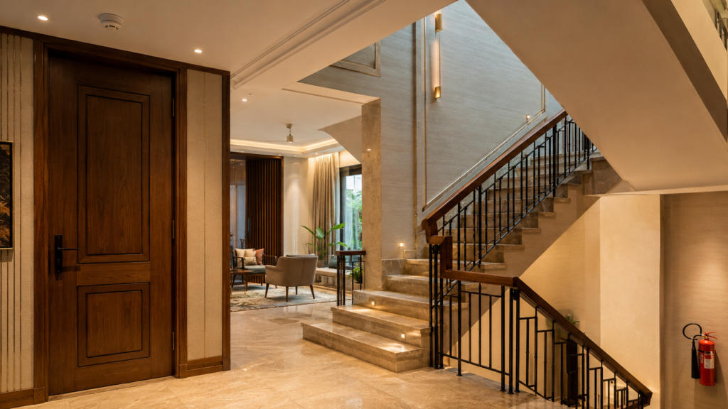 Hotel lobby with marble floor, warm lighting, a dark wood door on the left, and a grand staircase with black railing to the right.